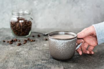 Latte in a ceramic cup in female hands and a jar of coffee beans on the table.