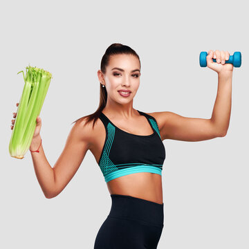 Square image. Smiling strong fitness woman wearing sport suit with dumbbell and celery on gray isolated background