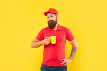 Serious bearded man in casual red cap and tshirt holding mug yellow background, coffee