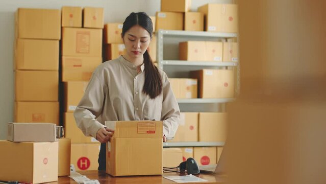 Asian woman working at online store warehouse packing product to parcel cardboard boxes, online e-commerce retail small business concept