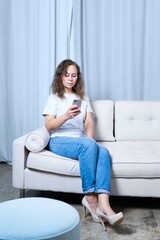 a girl in blue jeans and a white t-shirt looks at the phone while sitting on a light-colored sofa