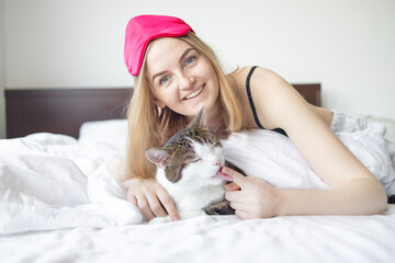 Young blonde 30s woman with sleep mask and cute cat lying on bed in the room at home. 