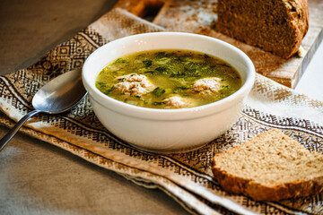 Homemade appetizing, delicious soup with meatballs, homemade bread, kitchen national napkin, and spoon on the kitchen dining table.