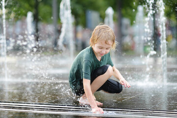 Little boy plays in the square between the water jets in the city fountain at sunny summer day....