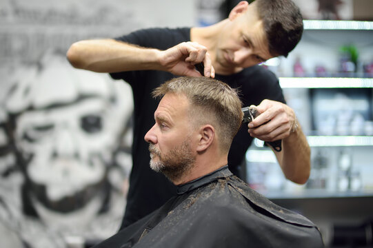 Hairdresser is cutting hair of handsome bearded mature man in salon. Stylist making hairstyle with electric shaver for person in barbershop. Services of a professional stylist. Fashion haircut