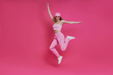 Full length of happy young woman in trendy clothing jumping against pink background