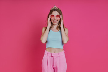 Happy young woman in funky crown looking at camera while standing against pink background