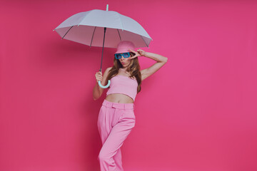 Playful young woman in trendy clothing holding umbrella while standing against pink background