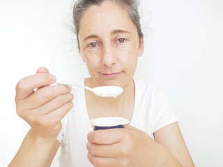 Forty nine year old woman in a white T-Shirt against a white background eating a youghurt