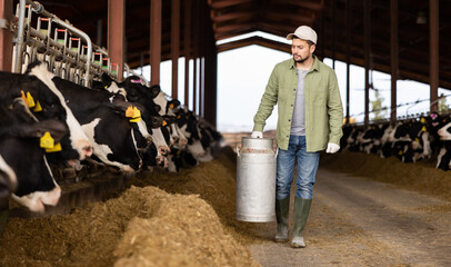 Focused young adult male farm worker carrying big milk can walking in cowshed on dairy farm