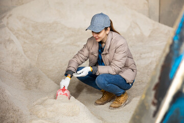 Woman farmer squatting at heap of corn flour in fodder storage and using scoop.