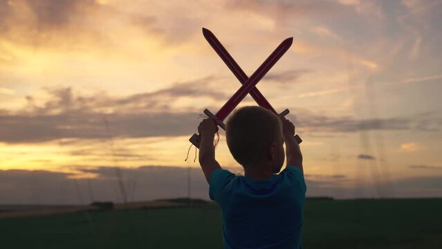 Child toy sword, children's dreams. Boy plays stands with swords raised in his hand, crossing them in sky, against background of sky, depicting medieval knight. Child boy playing superhero. Child Game