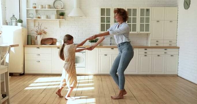 Happy small energetic kid daughter dancing barefoot on warm floor in modern kitchen with sincere mother. Emotional sincere two different generations family having fun, entertaining on weekend.