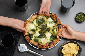 People taking slices of delicious pizza with pesto, cheese and basil at black table, top view