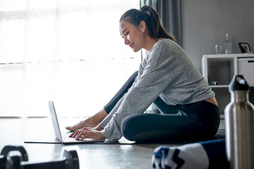 Sporty young Asian woman exercising at home, watching fitness video on Internet or having online fitness class, using laptop, living room interior, copy space