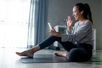 Sporty young Asian woman exercising at home, watching fitness video on Internet or having online fitness class, using smartphone, living room interior, copy space