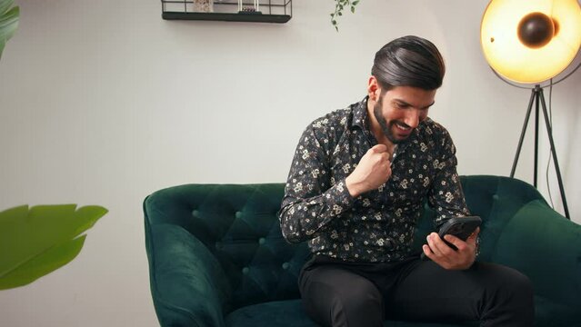 Latin handsome bearded man sitting on a green-colored couch looking at his mobile phone and cheering. Concept of victory. High quality 4k footage
