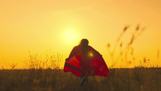 child superhero girl red cloak with long hair runs across green field sunset. chidhood dream red cloak flutters wind. game motion child. running girl superhero with long hair rays light concept dream