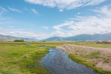 Scenic green landscape with mountain creek in sunlit grassy steppe among mountains under clouds in blue sky at changeable weather. Colorful scenery with mountain brook with clear water in bright sun. - obrazy, fototapety, plakaty