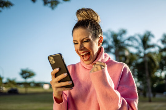 Image of excited young blond Latin woman making winner gesture using cellphone on park background