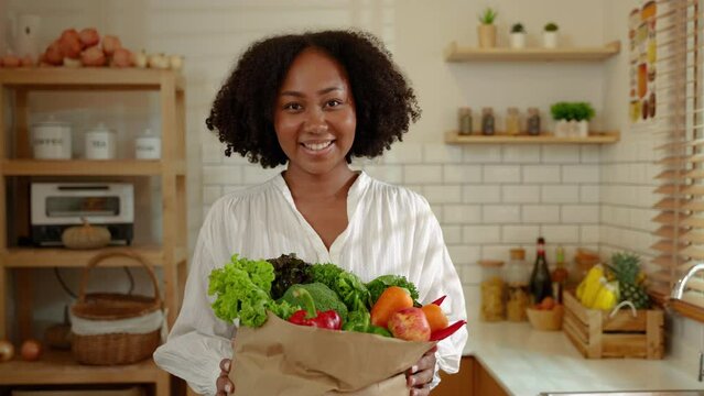 Smiling African American portrait woman happily holding a bag of vegetables in the kitchen, healthy eating and wellness concept.
