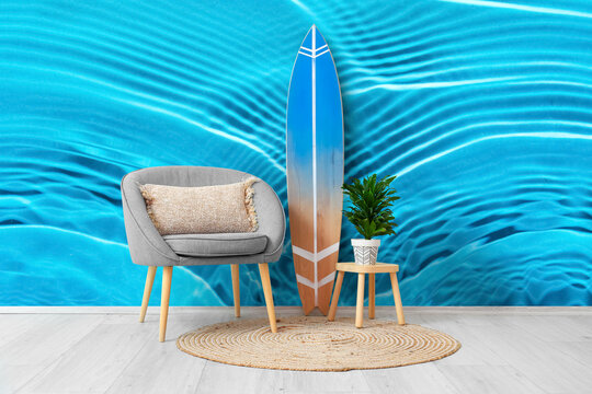 Armchair, stool and surfboard near wall with print of clear blue water