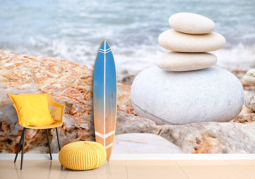 Surfboard, armchair and pouf near wall with print of pebbles on sea beach