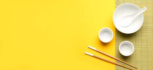 Chinese bowls with chopsticks and bamboo mat on yellow background with space for text