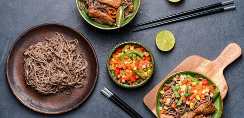 Plates with tasty soba noodles, vegetables and meat on dark background