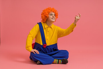 a cheerful clown in a wig and a yellow-blue suit sits cross-legged and calls someone with his finger on a colored background
