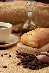 Rustic bread placed on a cutting board, around it a cup of American-style coffee, olive oil, coffee beans and garlic, placed in a vegetable fiber sack