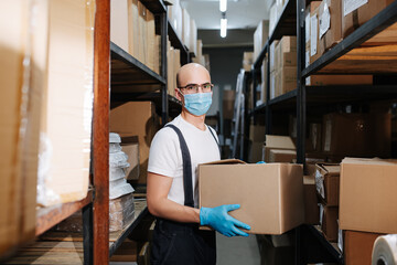 Masked male warehouse worker holding a box, hands in blue gloves