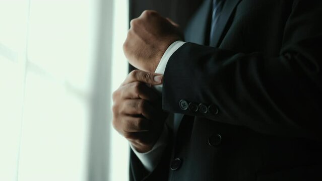 Confident businessman in classic blue suit buttoning or adjust cufflinks near window in hotel room at the morning. Handsome man wearing a nice suit on wedding day.