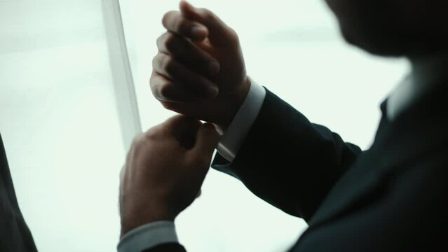 Confident businessman in classic blue suit buttoning or adjust cufflinks near window in hotel room at the morning. Handsome man wearing a nice suit on wedding day.