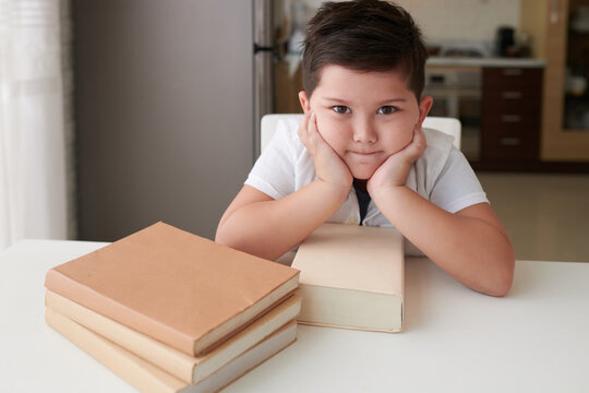 Smart determined little boy sitting at table with many books and looking at camera