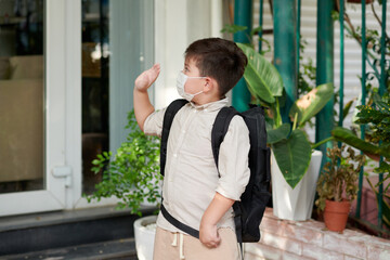 Fototapeta premium Schoolboy in medical mask waving with hand when leaving house for school