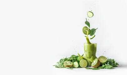 Green smoothie splashing in glass and flying ingredients: cucumber,kiwi and spinach leaves at white background with heap of green fruit and vegetable. Border. Front view.