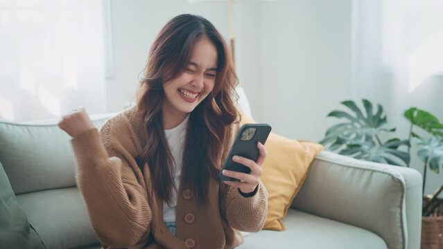 Excited young asian woman hold phone chatting in dating app feel happy sit on sofa at home, Closeup joyful female reading good news on phone in slow motion. Surprised lady celebrating victory on phone
