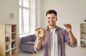 Portrait of happy young man with small layout of wooden house and with keys in hands. Handsome smiling guy who is buyer, homeowner or tenant of apartment shows keys while looking at camera. - obrazy, fototapety, plakaty