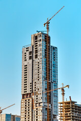 White and blue glass skyscraper under construction with a yellow tower crane along it and smaller...