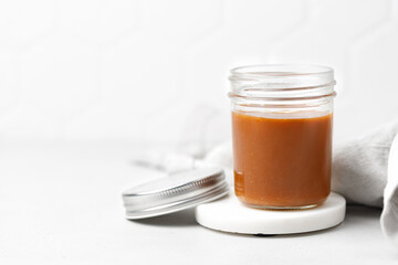 Salted caramel sauce in glass jar with lid on marble board, white background. Homemade caramel