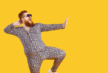 Fototapeta na wymiar Funny eccentric overweight man dressed in leopard print pajamas dancing and fooling around on orange background. Cheerful crazy bearded fat man in sunglasses having fun at copy space. Web banner.