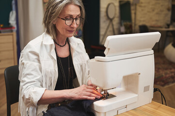 Mature European woman, fashion designer tailor sitting at wooden desk, using sewing machine sews clothes in a tailoring atelier