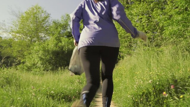 A young sporty woman running at the park and collects garbage. Back view. Follow shot. The concept of plogging and Earth Day.