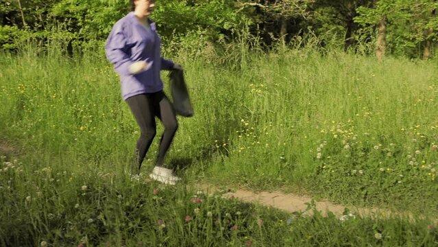 Plogging. A young woman jogging at the forest and pick up garbage. Side view. The concept of volunteering and environment protection.