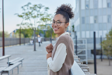 Outdoor shot of positive woman with curly hair smiles gladfully wears spectacles casual shirt and...