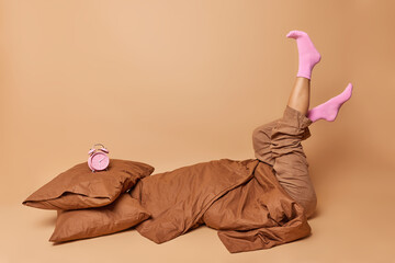 Indoor shot of unrecognizable woman dressed in pajama keeps legs raised up wears socks poses in bed under blanket doesnt want to get up isolated over beige studio background. Hard awakening.