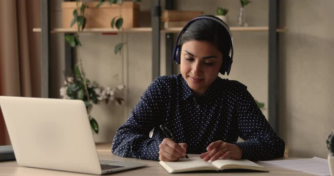 Video call event, self-education process use modern tech, internet connection concept. Indian student sit at desk listen audio course through headphones, taking notes writing down helpful information