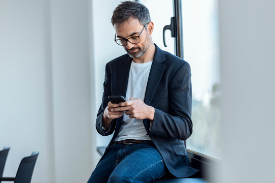 Businessman text messaging through smart phone sitting on window sill in office