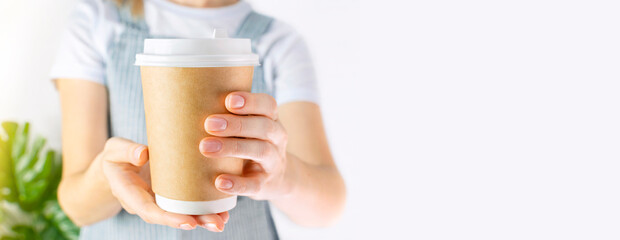 Unrecognizable young woman barista in an apron holds in her hands and gives a cardboard cup with a hot aromatic coffee drink or tea or cocoa. Female works in a coffee shop, coffee break. Copy space.